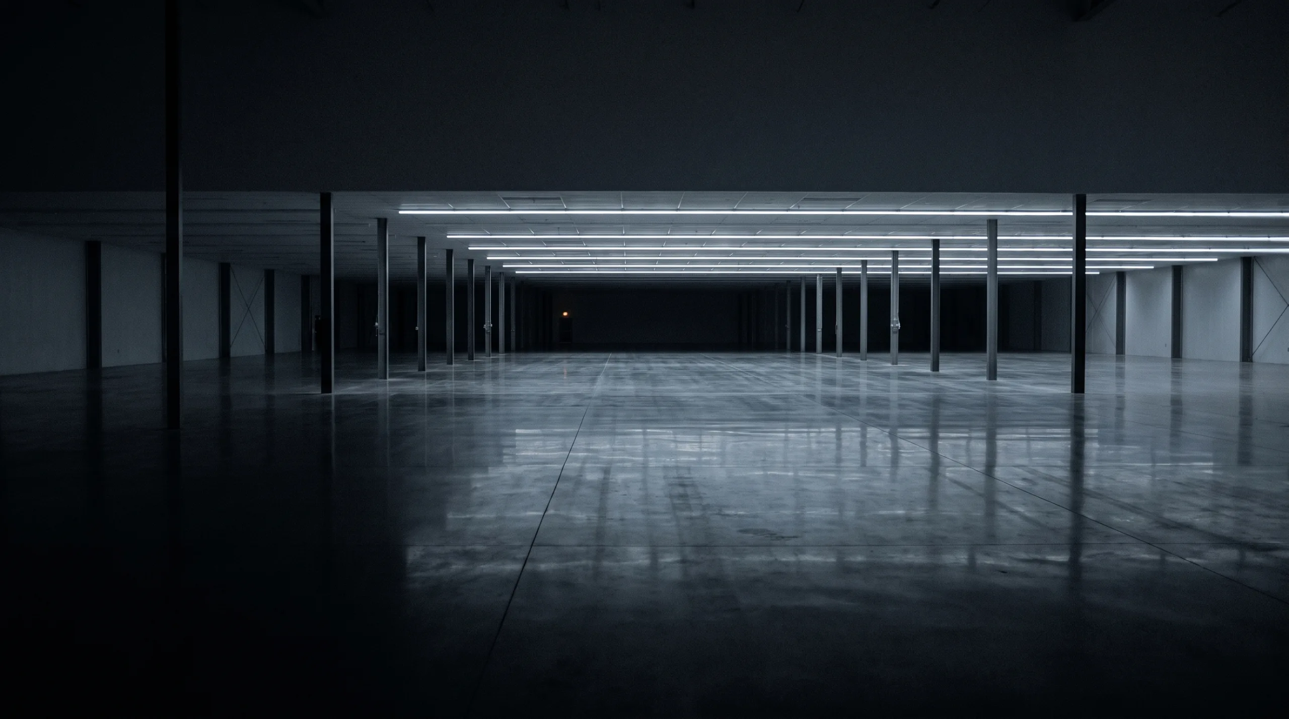 An empty industrial hall at night, lit only by receding ceiling light strips reflected in the floor.