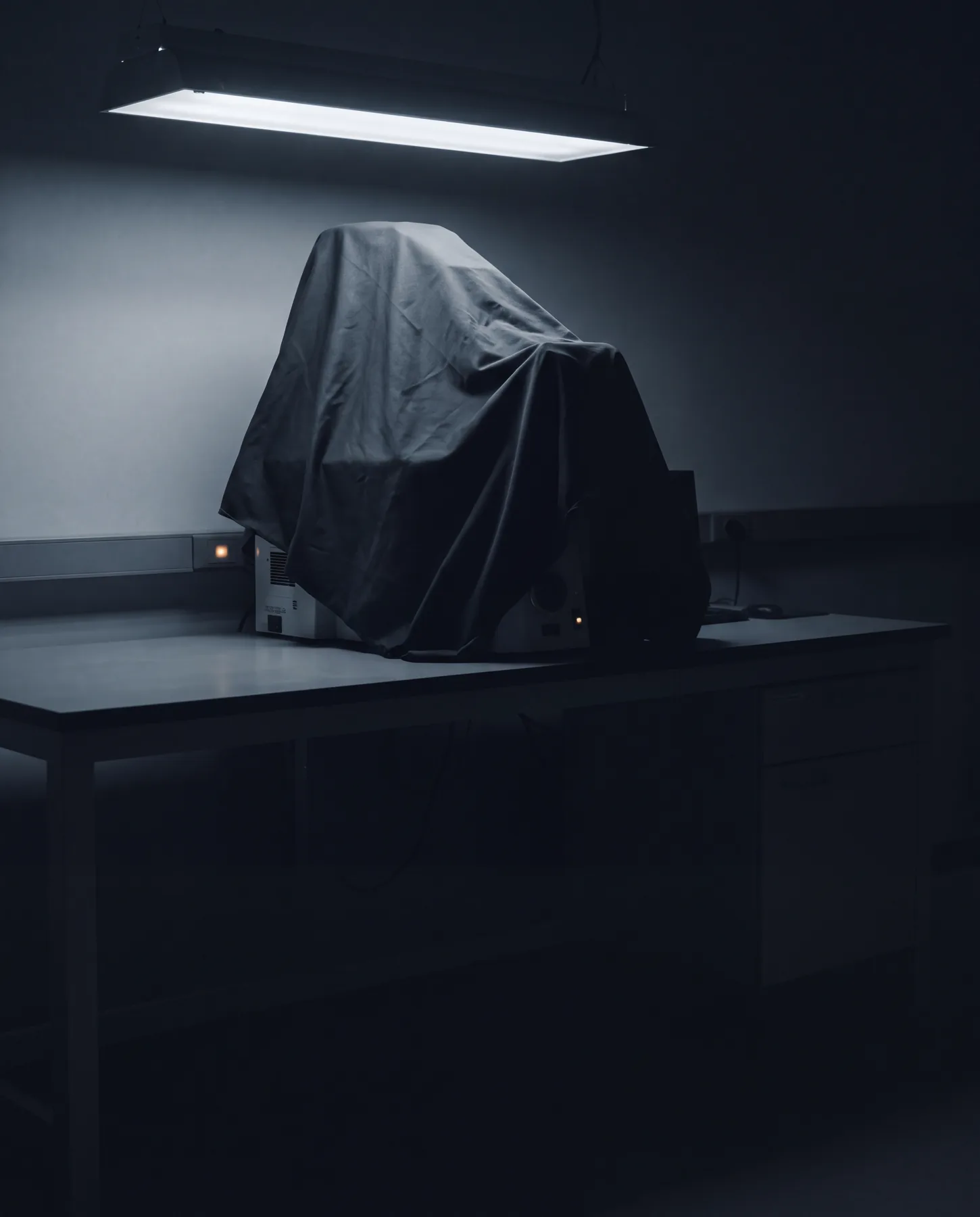A covered instrument on a laboratory bench beneath a single fluorescent tube.